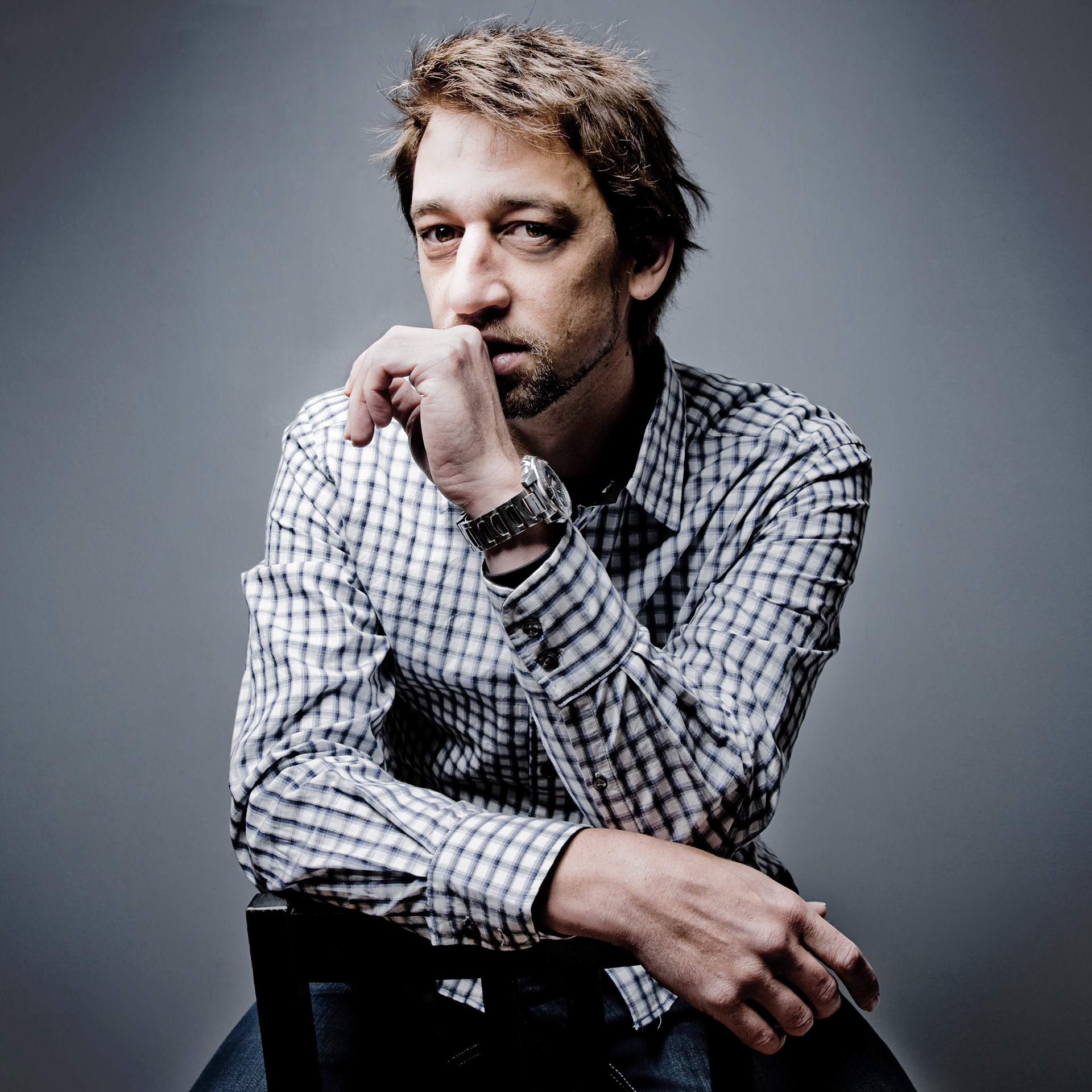 Studio portrait of a Marcos Appelt with tousled hair and a short beard wearing a checked shirt, seated with one hand resting near his face against a muted gray background, taken by Marcos Appelt Portrait Photographer in Bristol and Southwest.