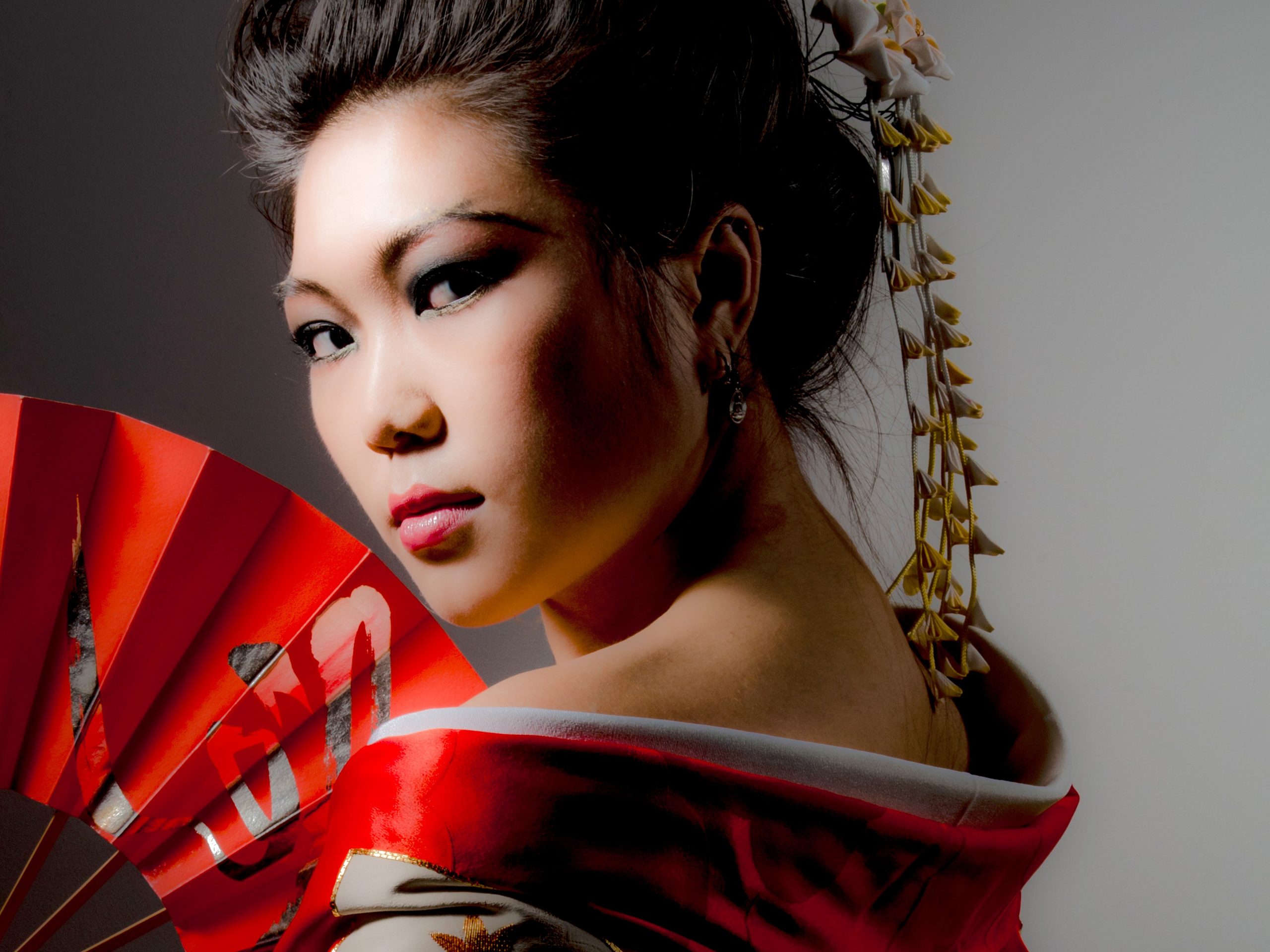 Studio portrait of a woman in stylized traditional attire holding a red fan, with dramatic makeup and lighting, posed in profile against a neutral background, taken by Marcos Appelt Portrait Photographer in Bristol and Southwest.