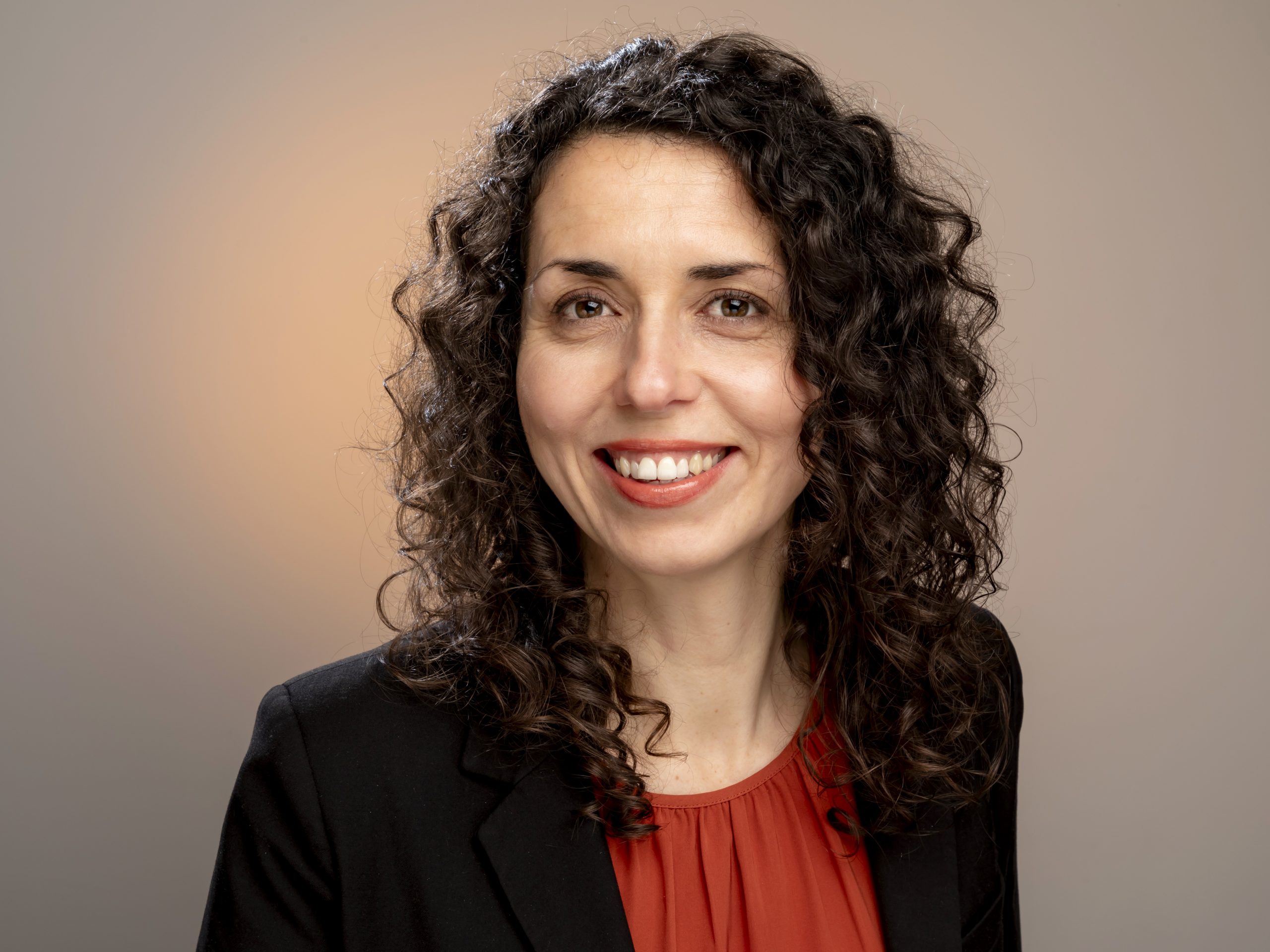 Studio portrait of a woman with long curly dark hair wearing a black blazer over an orange top, smiling softly against a warm gradient background, taken by Marcos Appelt Portrait Photographer in Bristol and Southwest.