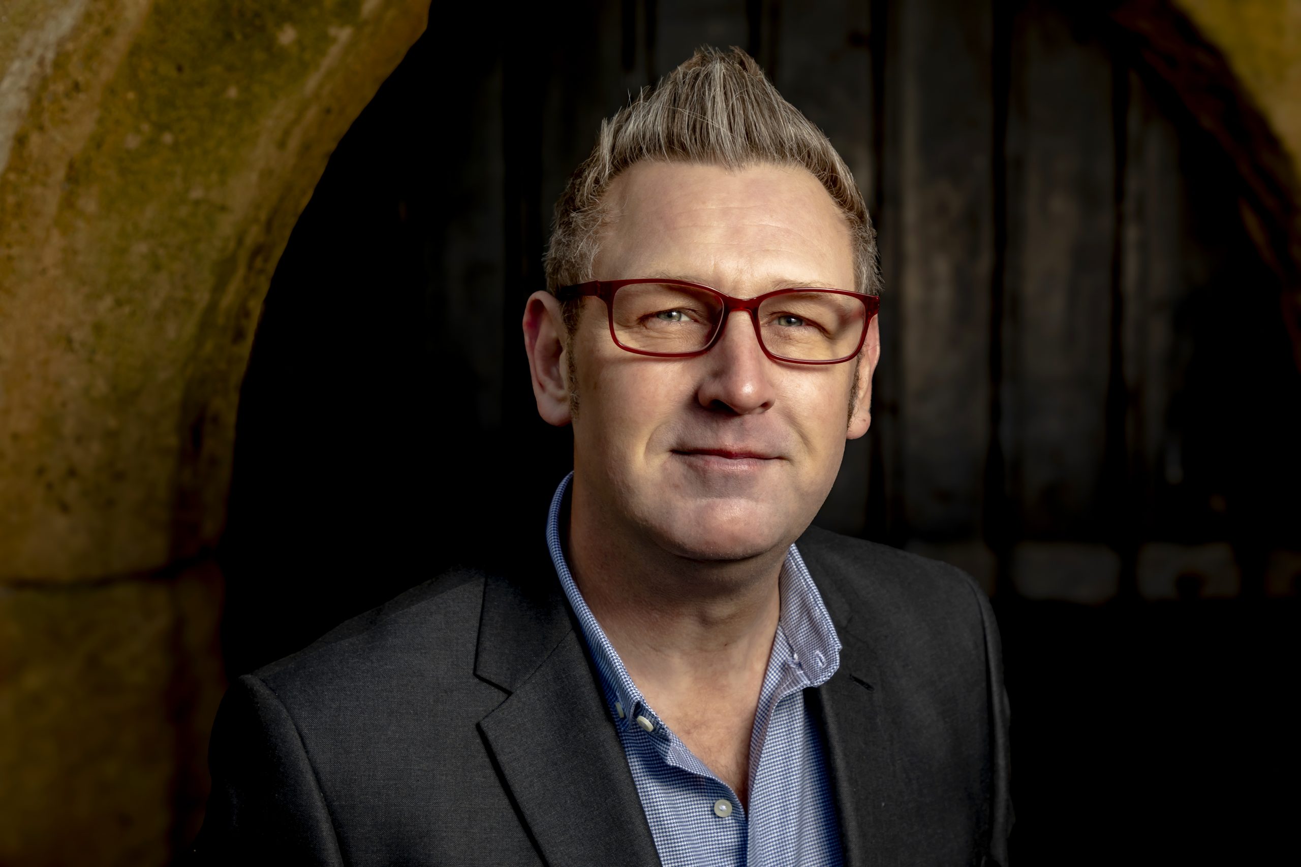 Professional headshot of a man wearing glasses and a suit jacket against a dark arched doorway, taken by Marcos Appelt Portrait Photographer in Bristol and Southwest.