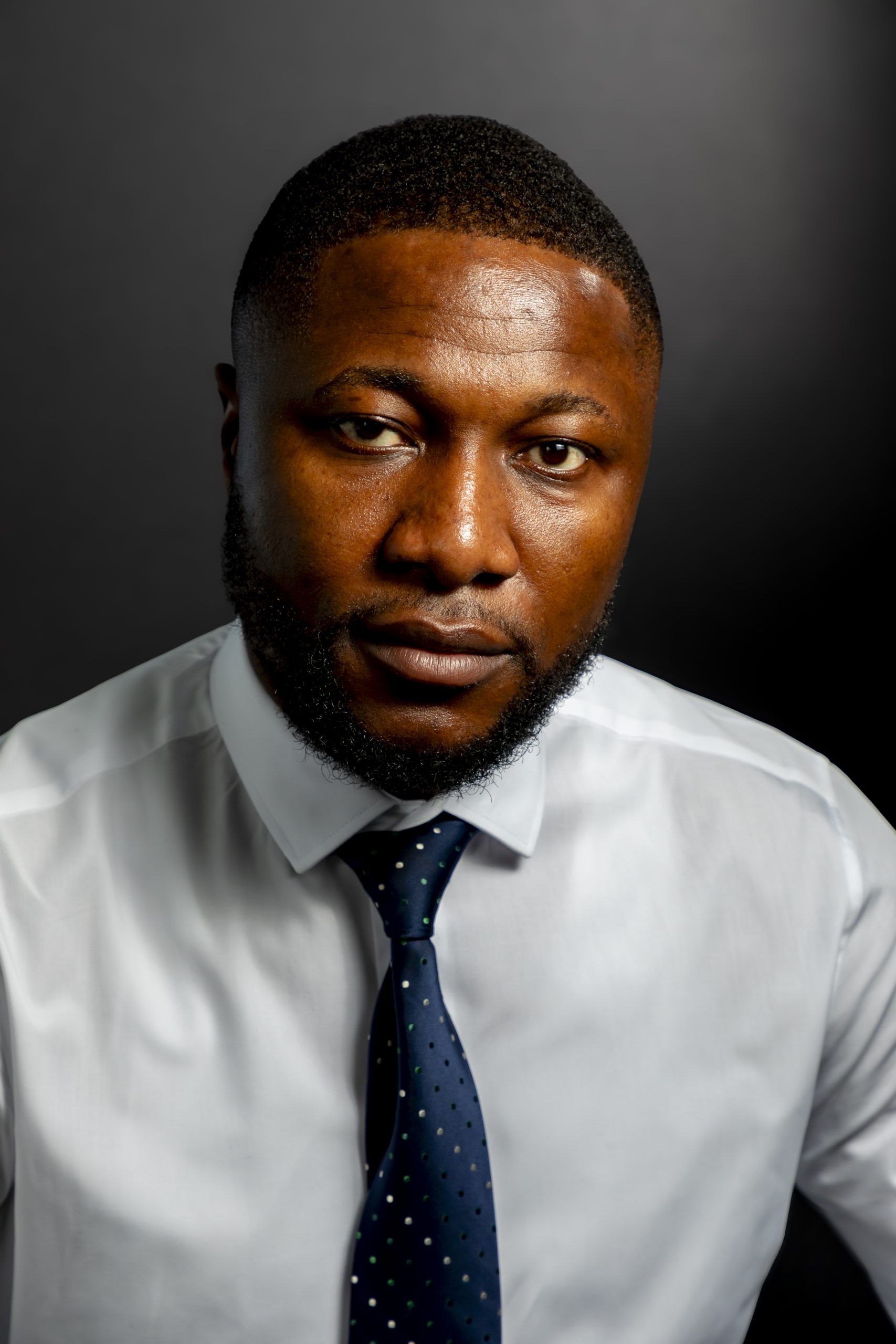 Studio portrait of a man with short hair and a full beard wearing a white dress shirt and a navy tie, lit dramatically against a dark grey background, taken by Marcos Appelt Portrait Photographer in Bristol and Southwest.